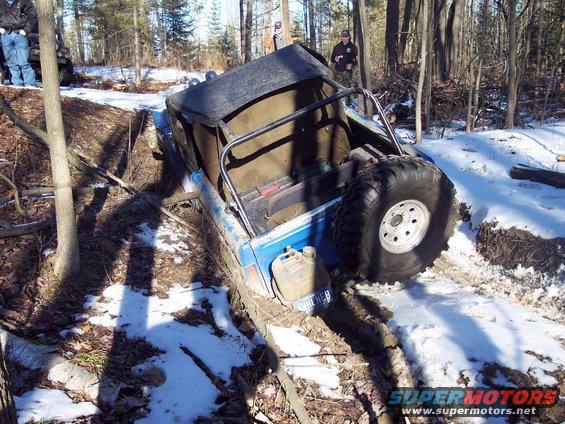 Blue off-road vehicle tilted in a muddy ditch with snow on the ground. Spare tire mounted on the back