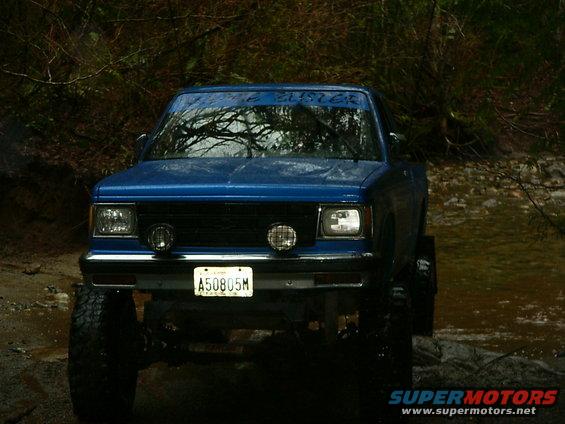 Blue pickup truck with large tires driving through water, trees in background. License plate A50805M