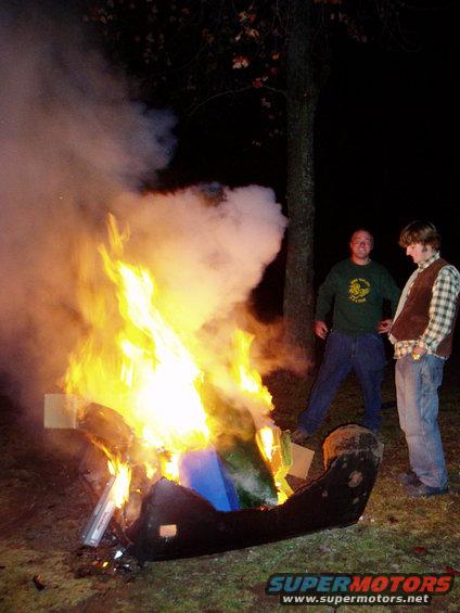 two guys watching a large bonfire with smoke and flames