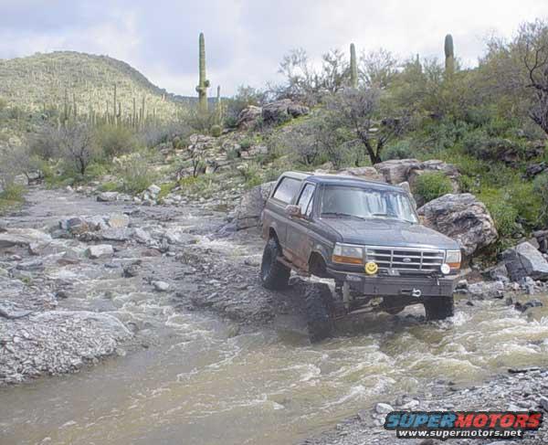 Dark gray Ford Bronco driving through a rocky stream in a desert landscape