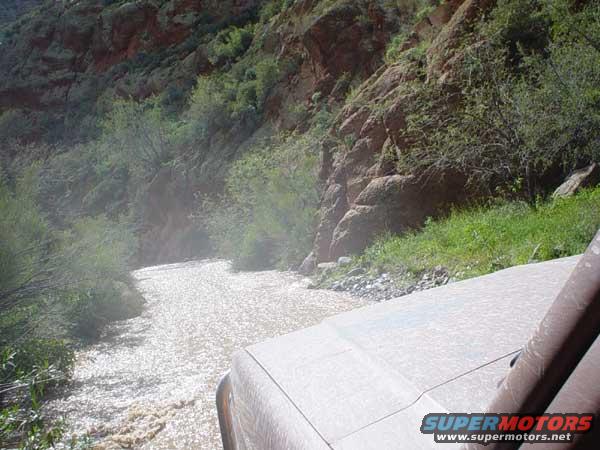 Vehicle driving through a shallow, rocky river in a canyon. Part of the vehicle's hood and windshield frame are visible