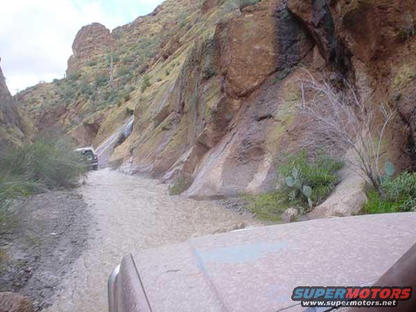 Two SUVs driving through a muddy canyon with a waterfall in the background