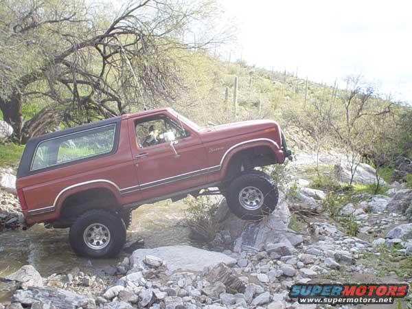 Red Ford Bronco with black top driving over rocks in a stream, desert landscape in background