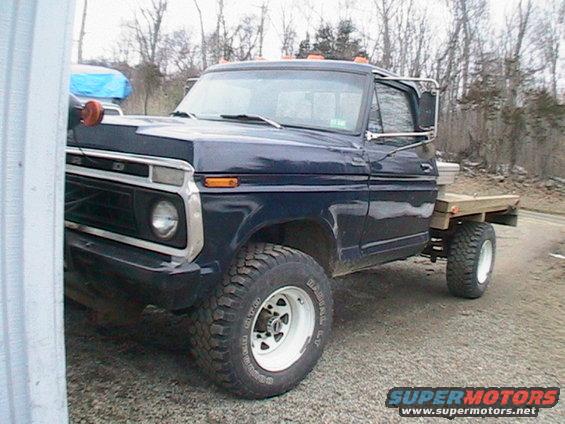 Dark blue 1977 Ford F-250 flatbed truck, front quarter view. It has large tires and a wooden bed
