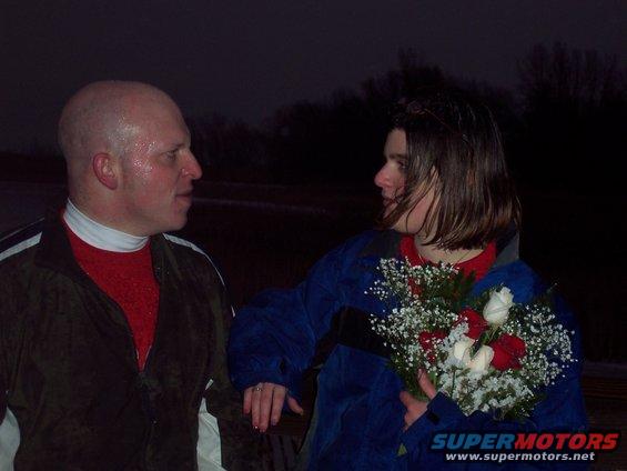 Bald man in red shirt and dark jacket talks to a woman holding a bouquet of red and white roses