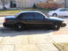1999 Ford Crown Victoria - A black sedan is parked on the street next to a sidewalk, with a white car visible in the background.
