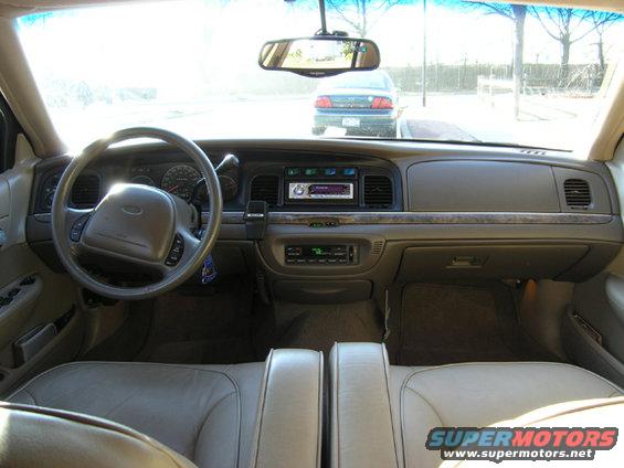 Car interior with tan leather seats, dashboard, steering wheel, and aftermarket radio. A blue car is visible through the windshield