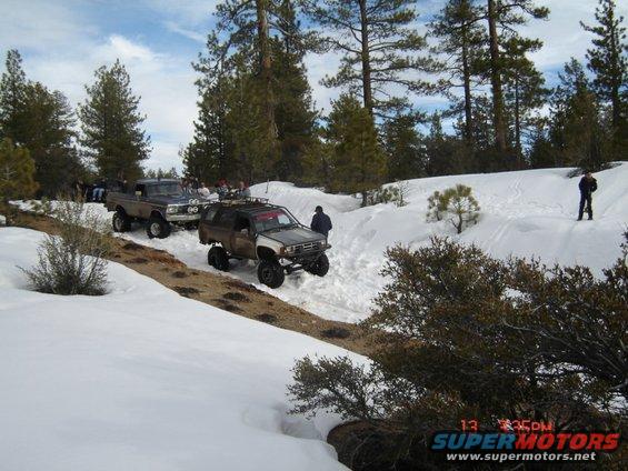 Two modified off-road vehicles driving on a snowy trail, one is a dark SUV with a roof rack, the other is a pickup truck with large headlights