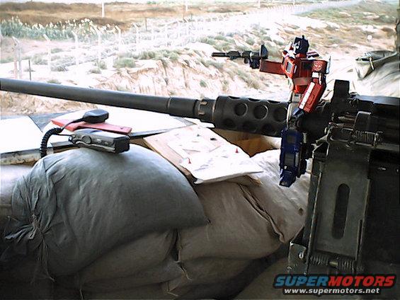 Optimus Prime toy standing on a large machine gun barrel, with sandbags and a radio in the foreground