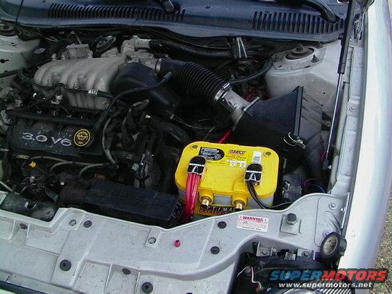 Engine bay with 3.0 V6 badge, yellow Optima battery, and Supermotors.net logo