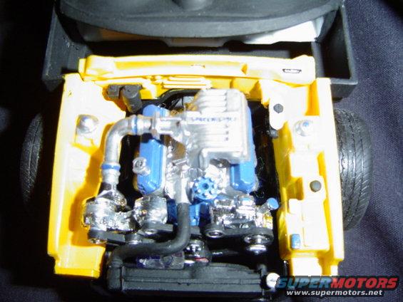Yellow model car engine bay with a silver and blue engine, chrome hoses, and black tires visible