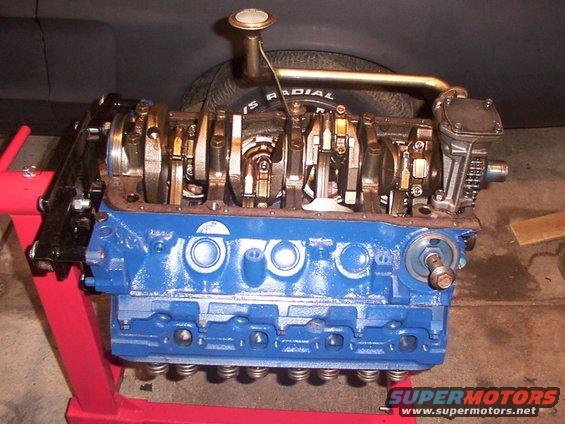 V8 engine block, blue, on red stand, with crankshaft visible. Tire in background