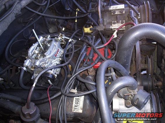 Engine bay showing a carburetor, red distributor cap, spark plug wires, and hoses. Part number FK408 visible on a black component