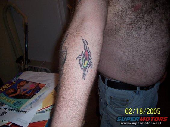 Tattoo of a ripped skin effect with colors and a 'G' on a hairy arm, next to a stack of books and a fan