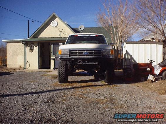 White Ford truck with lifted suspension and large tires, parked in a gravel driveway in front of a house