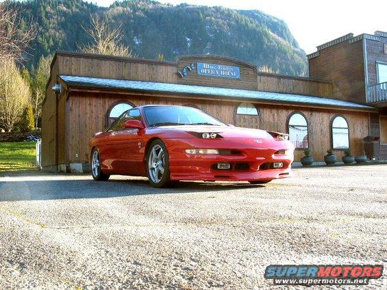 Red 1994 Ford Probe parked in front of a wooden building with mountains in the background