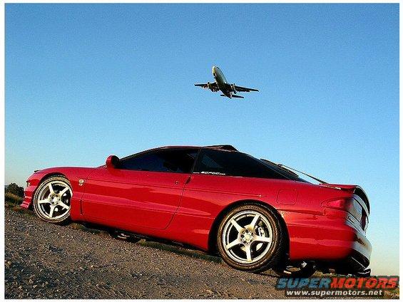 Red 1994 Ford Probe parked on a dirt incline with an airplane flying in the blue sky above
