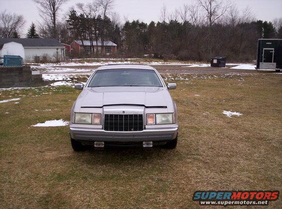 Front view of a silver sedan parked on grass with some snow patches. Buildings and trees in the background