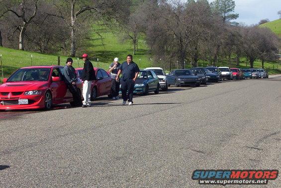 Several cars parked in a line on a road, with people standing near the front vehicles