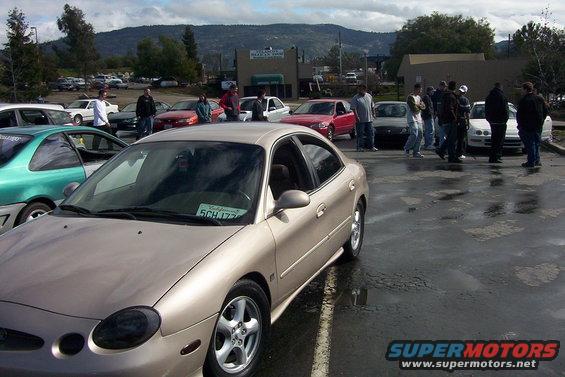 Gold sedan with a California plate parked in a lot with other cars and people