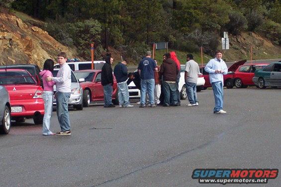 Group of people gathered around red and silver cars in a parking lot