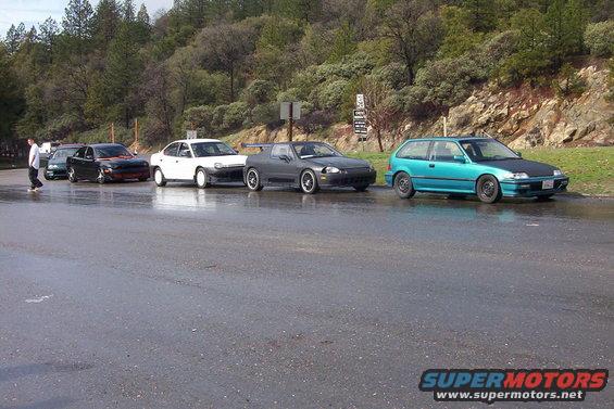 Several cars parked in a line on a wet road with trees and a hill in the background
