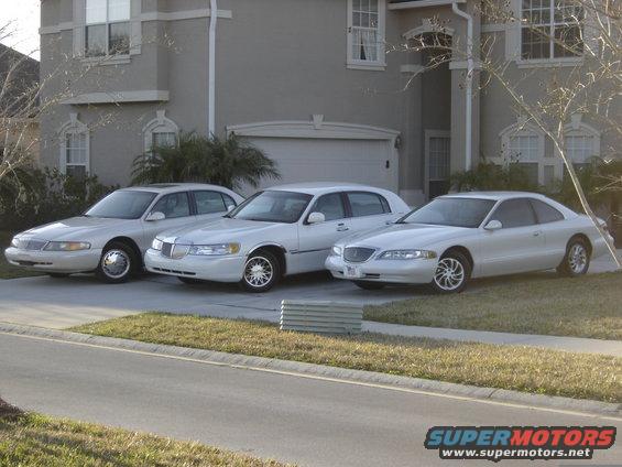 Three white Lincoln cars parked in front of a house. The car on the right is a Mark VIII