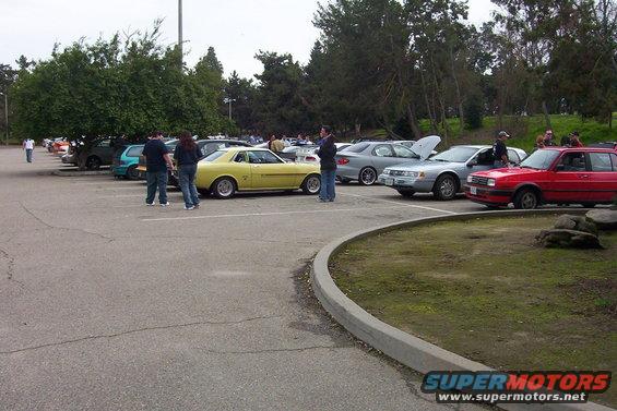 Yellow coupe parked in lot with several other cars, people standing around