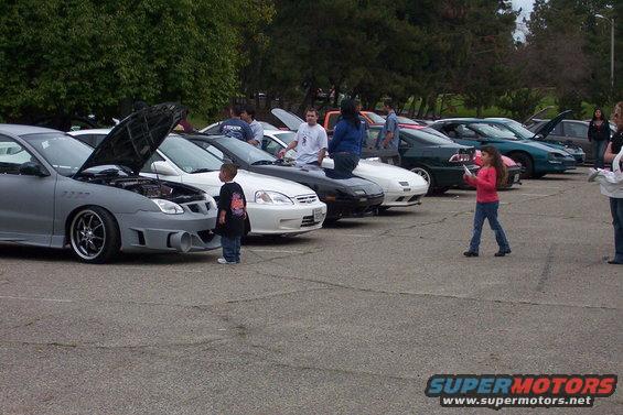 Several cars parked in a lot, some with hoods open, people standing around