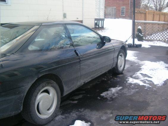 Dark coupe with a sloped rear window, seen from the side and slightly front. The car has a black door handle and a silver wheel cover