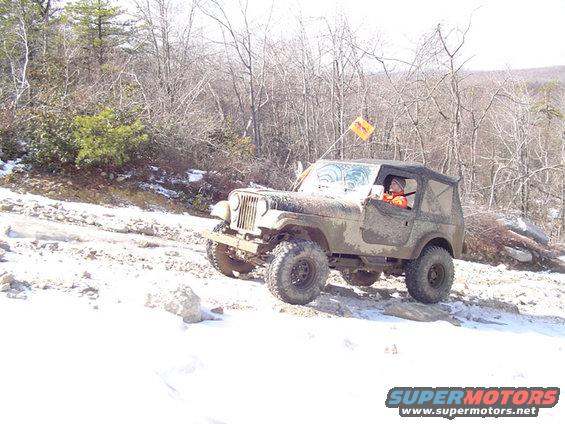 Muddy Jeep CJ-7 driving on a snowy, rocky trail with trees in the background