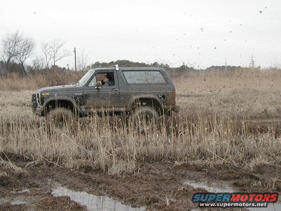 Dark colored Ford Bronco driving through a muddy field with tall dry grass. Mud is splashing