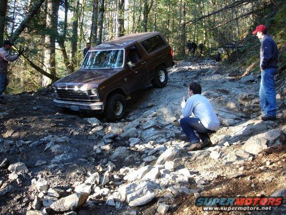 Brown 1978 Ford Bronco navigating a rocky trail in a forest with people watching