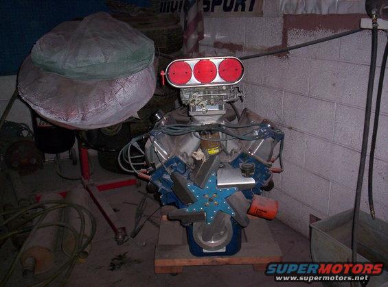 V8 engine with triple carb intake, blue fan, and orange oil filter. Engine is on a wooden stand