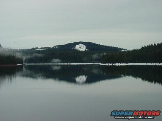 Calm lake reflecting forested hills with snow patches under an overcast sky. Watermark says SUPERMOTORS