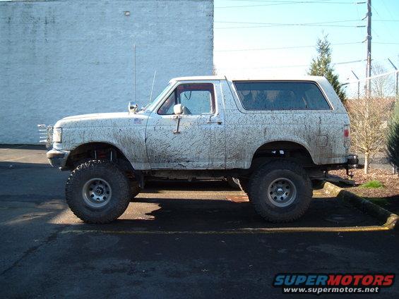 Lifted white Ford Bronco covered in mud, side view. Appears to be from the late 1980s