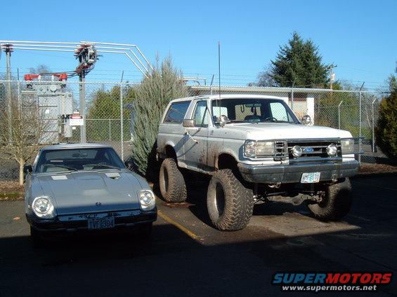 Lifted white Ford Bronco parked next to a silver Triumph TR7. Both vehicles are in a lot with a power substation in the background