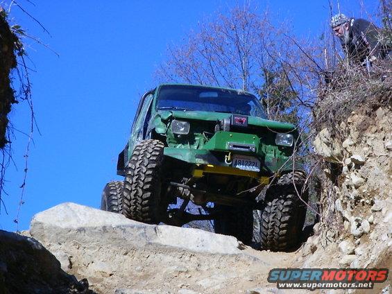 Green off-road SUV with large tires climbing over rocks, driver visible on right