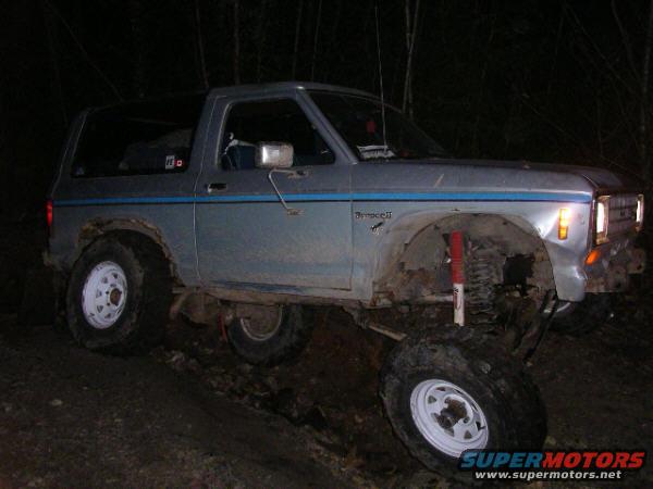 Gray 1984 Ford Bronco II with blue stripe, lifted suspension, and off-road tires on a dirt path at night