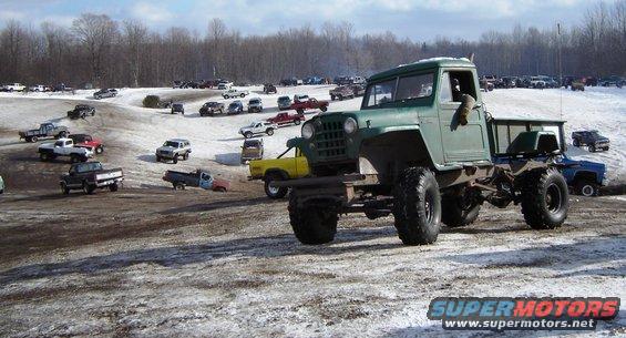 Green custom truck with big tires, parked in a snowy lot with other vehicles