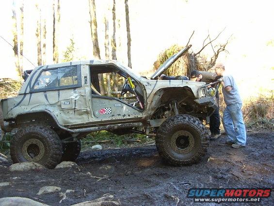 Heavily modified SUV, possibly a Jeep or similar off-road vehicle, with its hood open and two people attending to the engine bay. The vehicle is on a muddy, rocky trail