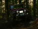 1990 Ford Bronco - A red Jeep CJ-7 navigates a challenging, muddy off-road course surrounded by dense forest.