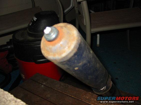 Rusty metal cylinder with blue paint and a bolt on top, angled over a wooden surface and red shop vac