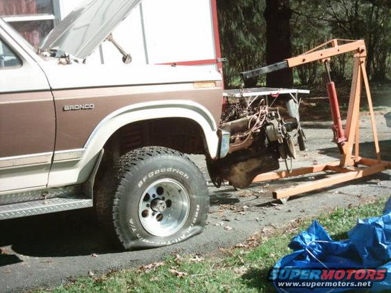 Ford Bronco with hood up and engine being removed by an engine hoist. Flat tire on front passenger side
