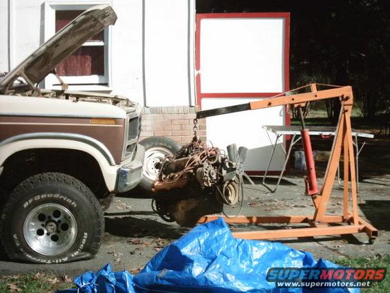 Brown and white 1980 Ford Bronco with hood open, flat front tire, and engine being removed by an orange engine hoist