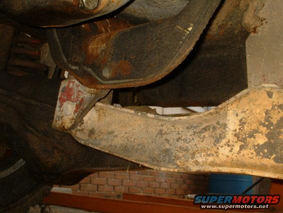 Underside of a vehicle, showing rusty suspension components and frame parts. A blue barrel is visible in the background