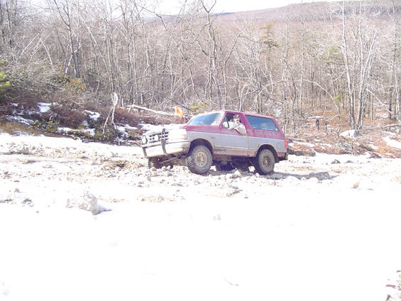 1995 Ford Bronco driving on a snowy, rocky trail with trees in the background