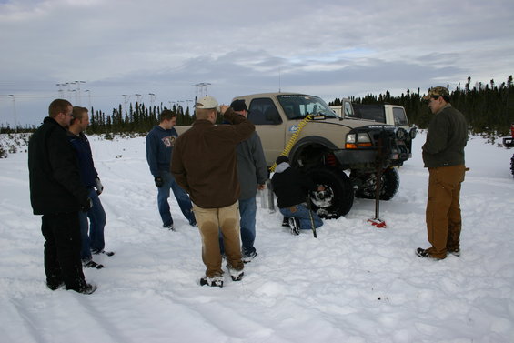 Tan pickup truck with a person changing a tire in the snow, with other people watching