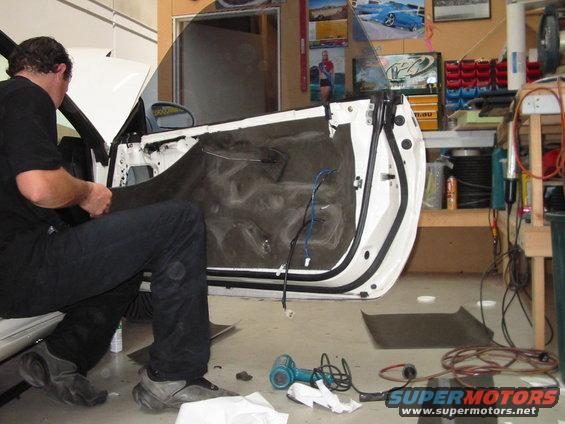 Man working on the interior of a car door, removing the panel. Tools and wires are visible on the floor