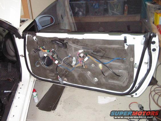 White car door interior with removed panel showing wiring, insulation, and speaker hole. A red spray can is on the floor